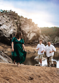 Mom, dad and toddler stand in a field with tall grass behind them with trees in the background. Mom is laying her head on dads shoulder and grabbing his arm wearing a black dress and tan coat. Dad is kissing moms head wearing a white sweater and gray jacket. Toddler is being held by dad with her hair in a ponytail wearing a tan coat.