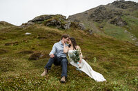 A couple sits together on a mossy hillside surrounded by rugged mountains during their Alaska elopement, sharing a quiet and intimate moment captured by Sydney Breann Photography.