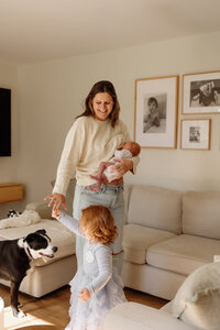A mother holds her newborn in her arm as she dances with her 2 year old in the living room. They are all wearing cream colored clothes. Captured with the finesse of a New England photographer, this moment exudes fine art elegance and documentary-style intimacy.