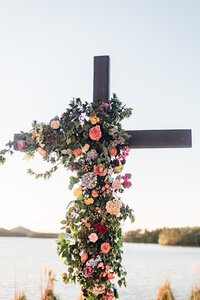 A cross covered in flowers and vines standing in front of water