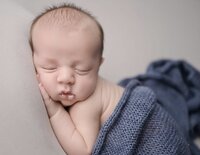 two week old baby boy sleeping posed with a blue knitted blanket over back.