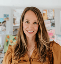 Stylish woman with long, highlighted hair and a warm smile, dressed in a ruffled mustard blouse, standing in a bright studio filled with vibrant artwork and photos pinned to the wall.