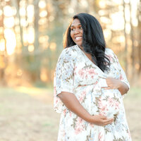 An expectant mother, wearing a white floral dress, smiles off to the side during her maternity photography session.