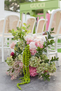 Lush pink and green floral arrangement beside rows of white cane-back chairs at an outdoor event with a branded backdrop.