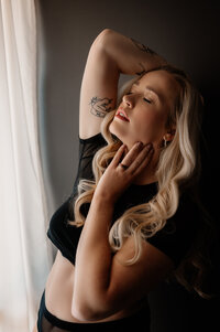 Boudoir portrait of blonde woman in black crop top, standing in front of a window with sheer curtains. Eyes closed and lips parted, she faces slightly off-camera with one hand in her hair and the other resting on her chin, creating a sensual and confident  look.
