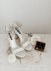 flatlay of bride's white heels, cream florals, and jewlery