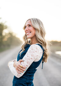 A high school senior smiling at the sky on a country dirt road. Her blonde hair is curled and she's wearing a denim dress layered with a white lace top underneath.