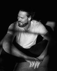 A black and white image of a bearded man in a white t-shirt sitting on the ground. His line of sight is looking the left of frame. He is sitting in a direct light.