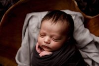 This is a detailed newborn photo captured by Mobile, AL newborn photographer. This newborn boy is wrapped in a dark grey swaddle, has his hand resting on his cheek, as he sleeps soundly in a brown bowl prop with light grey fabric in it for comfort.