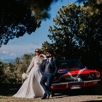 Coppia di sposi appoggiati ad una macchina d'epoca rossa.