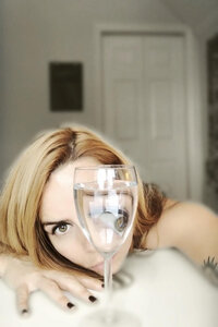 Artistic portrait of a woman peering through a wine glass filled with water, creating a distorted reflection of her eye and face.