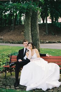bride and groom sitting on park bench, bride in white wedding dress, groom in black tie and tuxedo, wedding photographer