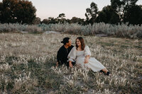 engaged couple sit in a field at golden hour for portraits