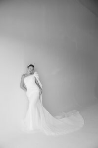 A woman in a wedding dress elegantly poses in a bright, white room, showcasing her joyful anticipation for the special day.