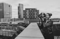 A person standing on a city rooftop with a camera up to their eyes