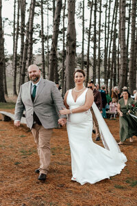 Bride and groom Dehlia and Nardus walking together after their outdoor forest wedding ceremony, with wedding party and guests celebrating in the background, photographed by Zanthe Vorsatz.