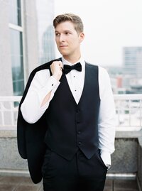 A groom stands for a portrait  holding his jacket over his shoulder looking off into the distance