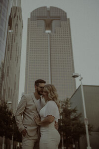 Editorial style downtown engagement session. Couple nuzzling together with an architecturally interesting building towering behind them.
