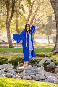 girl in blue cap and gown standing on a rock with her fist in the air