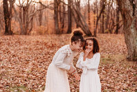 Two young sisters laughing together in a field of fallen leaves.  Both girls are wearing ivory dresses from Joyfolie.  Photo taken by Philadelphia family photographer, Kristi