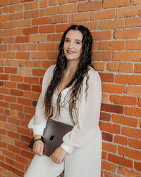 Taysha holding her closed laptop leaning against a red brick wall.