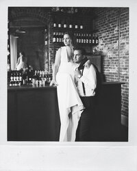 A bride sits on a bar in a polaroid with her groom snuggling her