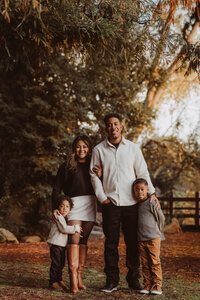 a family of four, mom and dad in the middle, two young boys on each side, as they stand in a park during the fall season 
