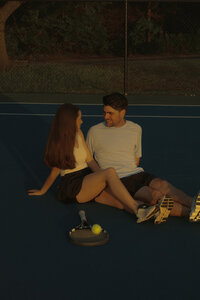 A man and woman sitting next to each other on a tennis court. They have rackets next to them  and are looking at each other as the sun lowers.