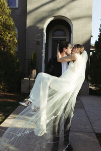 Couple embracing one another while sharing a kiss outside a grey building.