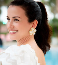 Close-up of a woman smiling poolside wearing Gaby & Grace handcrafted floral earrings made of white shells and delicate pearl accents. Styled with a white ruffled top for a refined, coastal look.