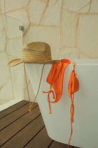 Straw beach hat and orange bikini draped over a white outdoor bathtub against a textured stone wall — sun-soaked and relaxed summer aesthetic.