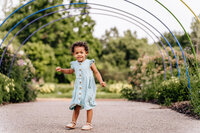 A young child in a light blue dress stands confidently under a series of colorful metal arches in a park, surrounded by greenery.