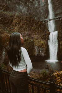 Rear view of woman standing at the base of a forest waterfall, wearing white – serene and wild brand photo by Mewphotovideo