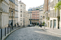 A cobblestone street lined with historic European buildings stretches downhill, reminiscent of charming Caribbean getaways. The architecture features balconies and ornate detailing. A few people stroll along the sidewalks on a clear day, adding a sense of liveliness to the scene.