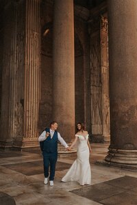 A couple holds hands while walking between the grand columns of an ancient building during their elopement in Rome, Italy. One partner wears a flowing white dress, while the other is dressed in a blue suit, both smiling as they enjoy their romantic moment.