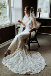 A bride in an elegant lace wedding gown with long train sits in a rattan chair by a window holding a champagne glass.