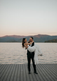 Groom lifting bride on a wooden dock at sunset with mountain views in the background, captured by a Spokane wedding photographer.