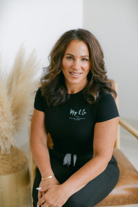A woman with long brown hair wearing a black outfit sits smiling on a brown chair with her hands clasped, next to a decorative arrangement of light-colored pampas grass at a Maine medical aesthetics clinic.