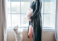 a dog standing on its back paws leaning against a windowsill and looking up at it's owner who is pregnant and she is holding her belly.