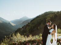 the groom is dipping the bride in front of the mountains. the sun is shining and the bride is smiling at the groom. the groom is looking directly at the bride. there are flowers in the background and the mountain is blue.