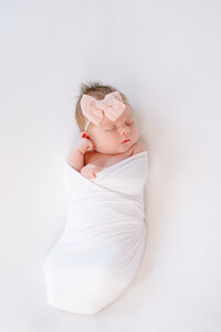a newborn baby girl is swaddled in a white wrap on a clean white background  with her hands up by her face while wearing a pink bow.  Image captured by Melissa Mayrie Photography of Charlotte, NC.