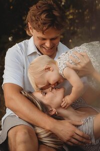 a mother laying in her husbands lap while she picks up her daughter and gives her a kiss