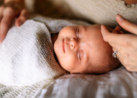 Close up shot of newborn babies face with a slight smile. She's wrapped in a white blanket and moms hand is gently touching her head.