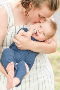 Mother holding her baby boy in her arms, kissing him on the head while the baby smiles at the camera.