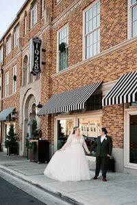 Waco wedding photographer captures lgbtq+ couple holding hands outside Hotel 1928, featuring classic striped awnings and historic brick architecture.Spring wedding reception tablescape on film photo at Ma Maison in Dripping Springs, Texas, featuring citrus inspired florals, pink napkins, and a view of the oak trees and fountain.