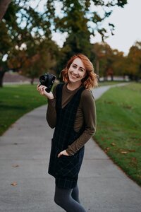 woman stands in middle of sidewalk smiling holding camera