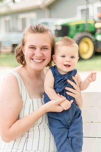 Mother sitting while her baby stands, holding hands and smiling at the camera.