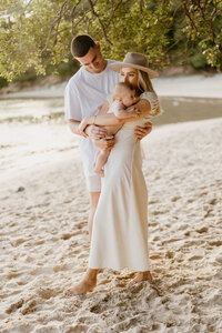 Couple embrace baby while standing on the beach - Natural family photoshoot