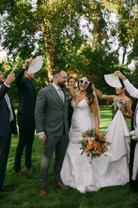 A groom in a green suit stands next to a bride in a white dress with heart shaped sunglasses being surrounded by family fanning them on a hot day