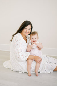 Mom in white dress sits with one year old baby girl in her lap during studio session with Worth Capturing